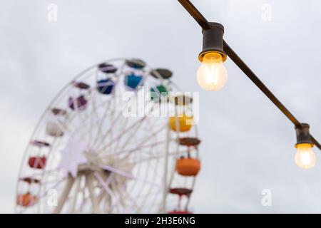 Lampade luminose con luci incandescenti appese al cielo nuvoloso e ruota panoramica colorata nel parco divertimenti sulla strada della città Foto Stock