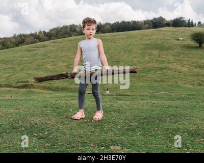 Corpo pieno di ragazzo serio guardando lontano mentre si sta in piedi con il fascio in mani su terreno erboso vicino a terreno collinare con alberi in campagna Foto Stock