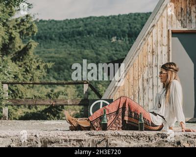 Vista laterale del corpo intero dell'hippie femminile in occhiali da sole seduti vicino a bevande alcoliche e struttura in legno contro la foresta verde in campagna Foto Stock