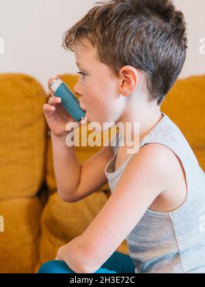 Vista laterale del ragazzino con asma usando l'inalatore mentre si siede sul divano a casa Foto Stock