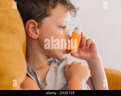 Vista laterale del ragazzino con asma usando l'inalatore mentre si siede sul divano a casa Foto Stock