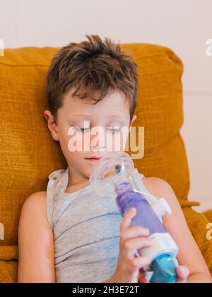 Contenuto ragazzo malato utilizzando respiratore e respirare l'ossigeno da inalatore mentre si siede sul divano a casa Foto Stock