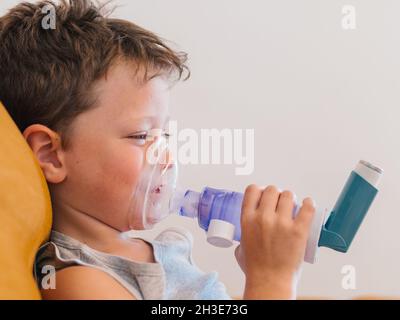 Vista laterale del contenuto ragazzo malato utilizzando respiratore e respirare ossigeno da inalatore mentre si siede sul divano a casa Foto Stock