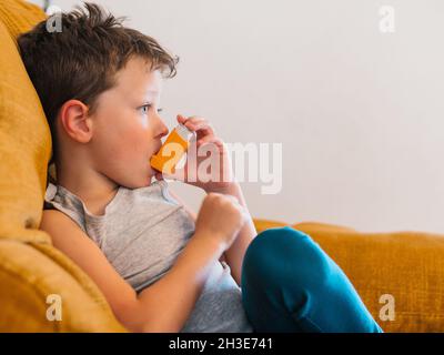 Vista laterale del contenuto ragazzo malato utilizzando respiratore e respirare ossigeno da inalatore mentre si siede sul divano a casa Foto Stock
