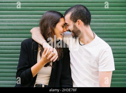 Allegra coppia di giovani ispanici in abiti casual ridendo con gli occhi chiusi mentre abbraccia e tocca la fronte vicino al muro verde sulla stree della città Foto Stock