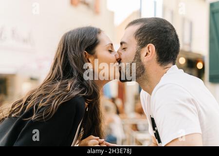 Vista laterale della giovane coppia etnica positiva baciare con gli occhi chiusi seduti in strada caffè in giorno di sole Foto Stock
