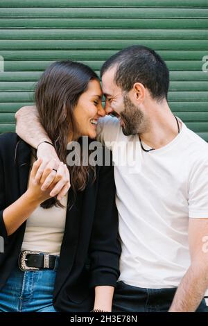 Allegra coppia di giovani ispanici in abiti casual ridendo con gli occhi chiusi mentre abbraccia e tocca la fronte vicino al muro verde sulla stree della città Foto Stock