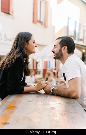 Vista laterale della romantica giovane coppia ispanica che tiene le mani e si guarda felicemente mentre si siede al tavolo in strada caffè Foto Stock