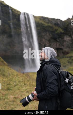 Vista laterale del fotografo turistico maschile in caldi outerwear e zaino ammirando la vista incredibile della cascata Seljalandsfoss che scorre attraverso la scogliera rocciosa in Foto Stock