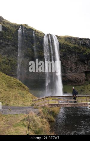 Vista laterale di turista maschile irriconoscibile in caldo outerwear e zaino in piedi su ponte di legno e ammirando la vista incredibile di Seljalandsfoss Wate Foto Stock