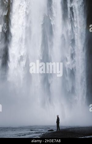 Vista laterale dell'escursionista maschile in abiti casual in piedi sul fiume sotto la spettacolare cascata di Skogafoss che scorre attraverso la massiccia scogliera rocciosa coperta con Foto Stock