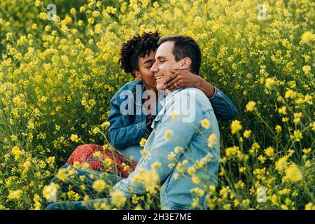 Coppia giovane amorevole multirazziale in abiti casual baciare mentre si siede in lussureggiante prato fiorente durante la data romantica il giorno di sole Foto Stock