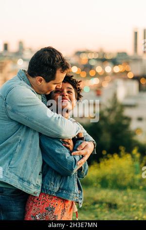 Coppia romantica e variegata che si abbraccia e si guarda l'un l'altro mentre si alza sul prato contro il paesaggio urbano con edifici su sfondo sfocato Foto Stock