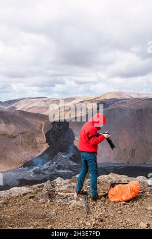 Vista laterale del fotografo maschile in outerwear in piedi sulla cima di una scogliera rocciosa vicino al vulcano attivo Fagradersfjall con lava nera in Islanda durante il giorno Foto Stock