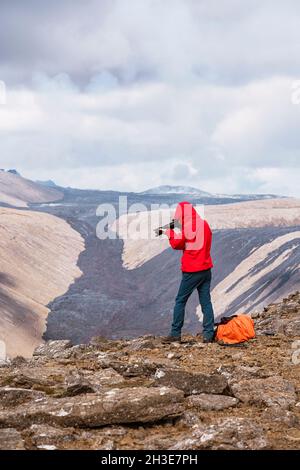 Vista laterale del fotografo maschile in outerwear in piedi sulla cima di una scogliera rocciosa vicino al vulcano attivo Fagradersfjall con lava nera in Islanda durante il giorno Foto Stock