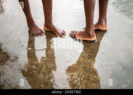 Da sopra di raccolto irriconoscibile a piedi nudi nero persone in piedi in piccola pozzanghera su strada asfaltata su São Tomé e Príncipe isola alla luce del giorno Foto Stock