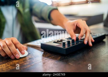 Ritagliare un musicista anonimo che lavora sul computer al tavolo mentre sintonizza il suono del controller MIDI nella stanza della luce Foto Stock