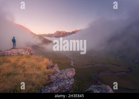 Vista posteriore di lontano irriconoscibile turista in piedi su terreno erboso circondato da montagne ruvide nella natura della Spagna in tempo nebbroso Foto Stock