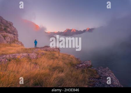 Vista posteriore di lontano irriconoscibile turista in piedi su terreno erboso circondato da montagne ruvide nella natura della Spagna in tempo nebbroso Foto Stock