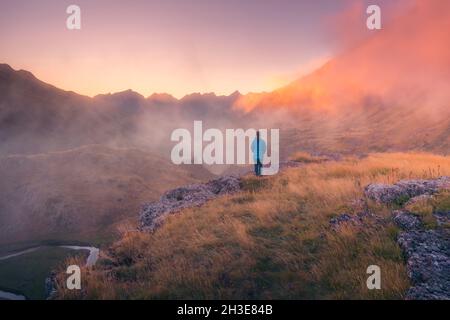 Vista posteriore di lontano irriconoscibile turista in piedi su terreno erboso circondato da montagne ruvide nella natura della Spagna in tempo nebbroso Foto Stock