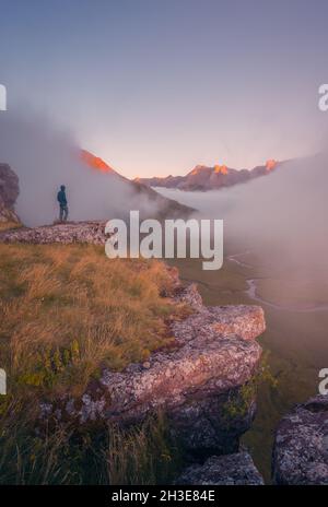 Vista posteriore di lontano irriconoscibile turista in piedi su terreno erboso circondato da montagne ruvide nella natura della Spagna in tempo nebbroso Foto Stock