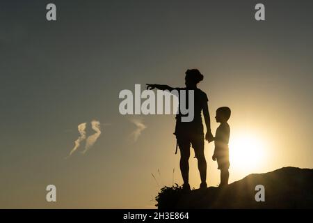 Madre e figlio contro la luce che si staglia dal sole puntando con le braccia verso l'orizzonte al tramonto Foto Stock