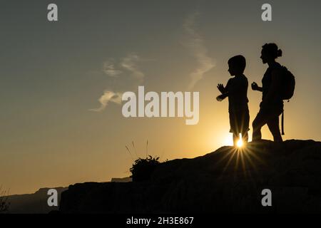 Madre e figlio contro la luce che si staglia dal sole puntando con le braccia verso l'orizzonte al tramonto Foto Stock