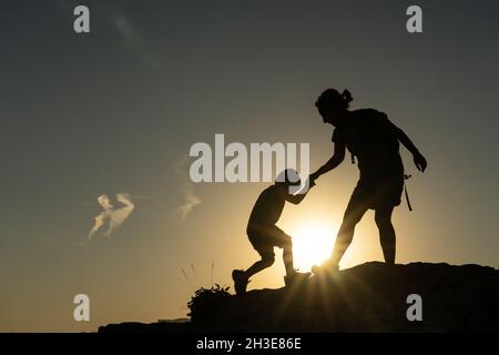 Madre che aiuta suo figlio a scalare una montagna contro la luce che il sole ha in una storia motivazionale Foto Stock