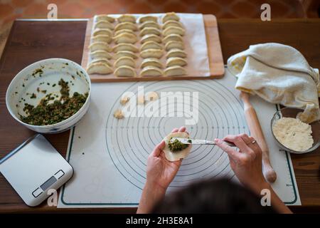 Dall'alto di raccolto irriconoscibile Donna in abiti casual e grembiule ripieno gnocchi con carne mentre preparando tradizionale cinese jiaozi in cucina Foto Stock