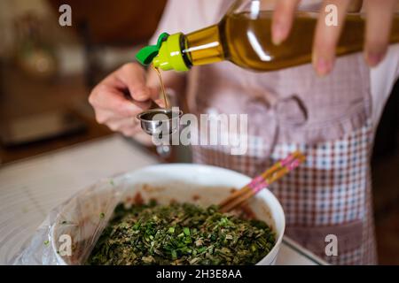 Da sopra di raccolto non riconoscibile casalinga in grembiule versando l'olio di oliva in ciotola con carne tritata cruda ed erbe mentre preparano il ripieno per jiaozi du Foto Stock