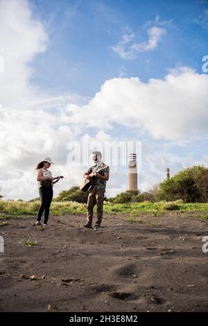 Corpo pieno di musicista maschile che suona chitarra acustica e guarda un amico femminile che suona ukulele mentre in piedi in natura in giorno di sole Foto Stock