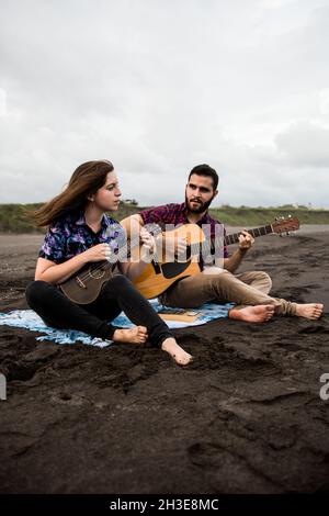 L'intera lunghezza di uomo sorridente suona la chitarra acustica con un amico positivo che suona l'ukulele mentre si siede sulla costa sabbiosa in natura in una giornata nuvolosa Foto Stock