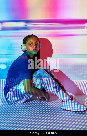 Vista laterale della giovane donna domenicana alla moda millennial con lunghe trecce afro seduti sul pavimento e guardando lontano mentre si ascolta la musica in headpho Foto Stock