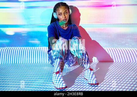 Vista laterale della giovane donna domenicana alla moda millennial con lunghe trecce afro seduti sul pavimento e guardando lontano mentre si ascolta la musica in headpho Foto Stock