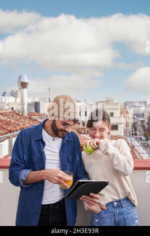 Giovane maschio con bicchiere di succo d'arancia in piedi vicino femmina flatmate mangiare sana mela verde sul tetto e navigare tablet insieme Foto Stock