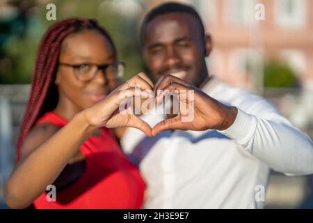 Felice coppia afroamericana abbracciando e mostrando segno a forma di cuore con le mani mentre si alza in strada in giorno di sole Foto Stock
