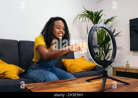 Donna afroamericana in abiti denim sorridente mentre si siede sul divano e toccando smartphone su una lampada ad anello incandescente Foto Stock