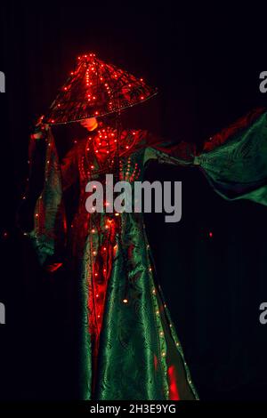 Donna enigmatica irriconoscibile in abito tradizionale creativo e capo vietnamita con illuminazione rossa in piedi in studio scuro su backgrou nero Foto Stock