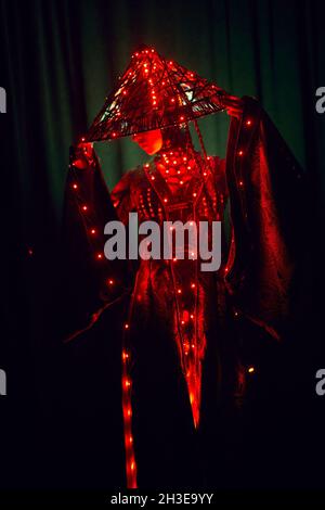 Donna enigmatica irriconoscibile in abito tradizionale creativo e capo vietnamita con illuminazione rossa in piedi in studio scuro su backgrou nero Foto Stock