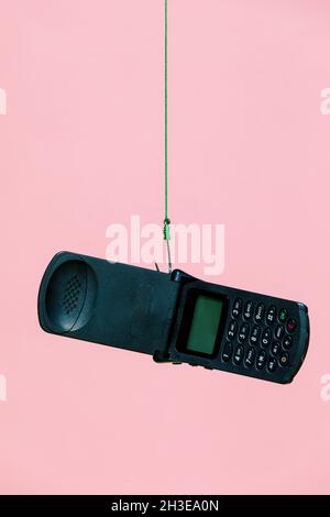 Vecchio telefono cellulare nero aperto flip appeso su gancio di metallo con corda verde su sfondo rosa in studio moderno chiaro Foto Stock