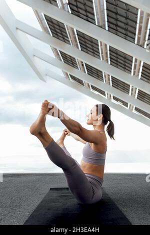 Vista laterale del corpo della donna sportiva in abbigliamento sportivo praticando Ubhaya Padangushthasana postura vicino al moderno pannello fotovoltaico durante l'allenamento yoga su s. Foto Stock