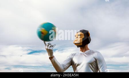 Persona irriconoscibile che indossa la maschera geometrica della scimmia che gira la palla in forma di Terra contro il cielo blu nuvoloso Foto Stock