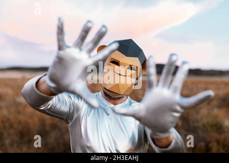 Persona anonima che indossa una maschera di scimmia geometrica mentre dimostra le mani con i pollici piegati alla macchina fotografica contro sfondo sfocato della campagna Foto Stock