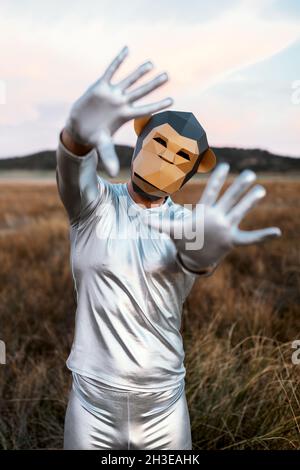 Persona anonima che indossa una maschera di scimmia geometrica mentre dimostra le mani con i pollici piegati alla macchina fotografica contro sfondo sfocato della campagna Foto Stock