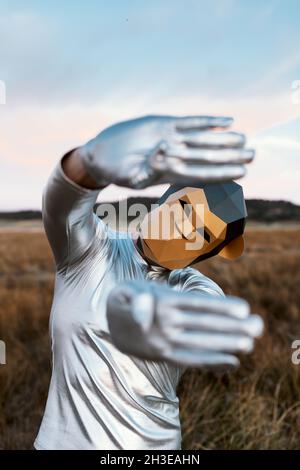 Persona anonima che indossa una maschera di scimmia geometrica mentre dimostra le mani con i pollici piegati alla macchina fotografica contro sfondo sfocato della campagna Foto Stock