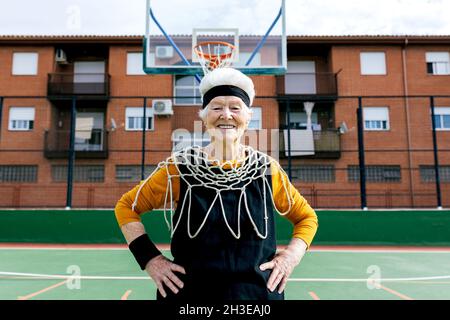 Donna matura sorridente in abbigliamento sportivo e rete bianca che guarda la macchina fotografica mentre si trova in piedi sul terreno sportivo con basket hoop durante l'allenamento Foto Stock