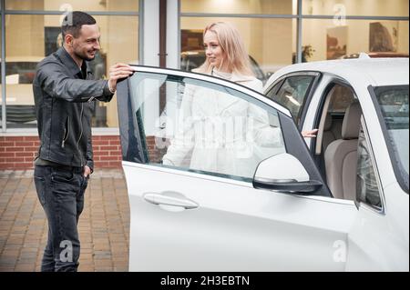 Bella giovane donna che sceglie auto in concessionaria all'aperto, parlando con bell'uomo. Foto Stock