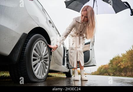 Bella donna su una strada ha fermato la sua auto bianca con pneumatico forato auto. Donna conducente che cerca di sgonfiare lo pneumatico, tenendo l'ombrello. Foto Stock