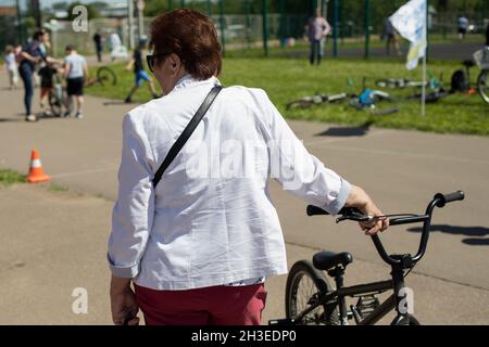 Donna anziana in giacca bianca con bicicletta. Giorno di sole, parco cittadino. Concorsi. Stile di vita della città Foto Stock