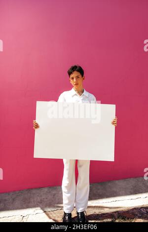 Ragazzo assertivo che tiene un cartello bianco su sfondo rosa. Ragazzo adolescente fiducioso che mostra un banner vuoto in uno studio. Giovane ragazzo gay comi Foto Stock